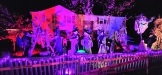 A house lit pink and purple at night with a row of life-size witch figures and a glowing cauldron behind a white picket fence
