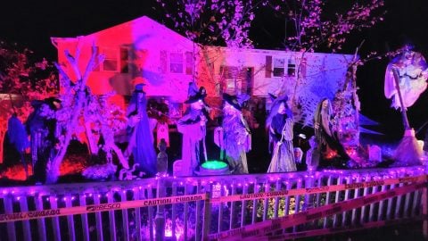 A house lit pink and purple at night with a row of life-size witch figures and a glowing cauldron behind a white picket fence