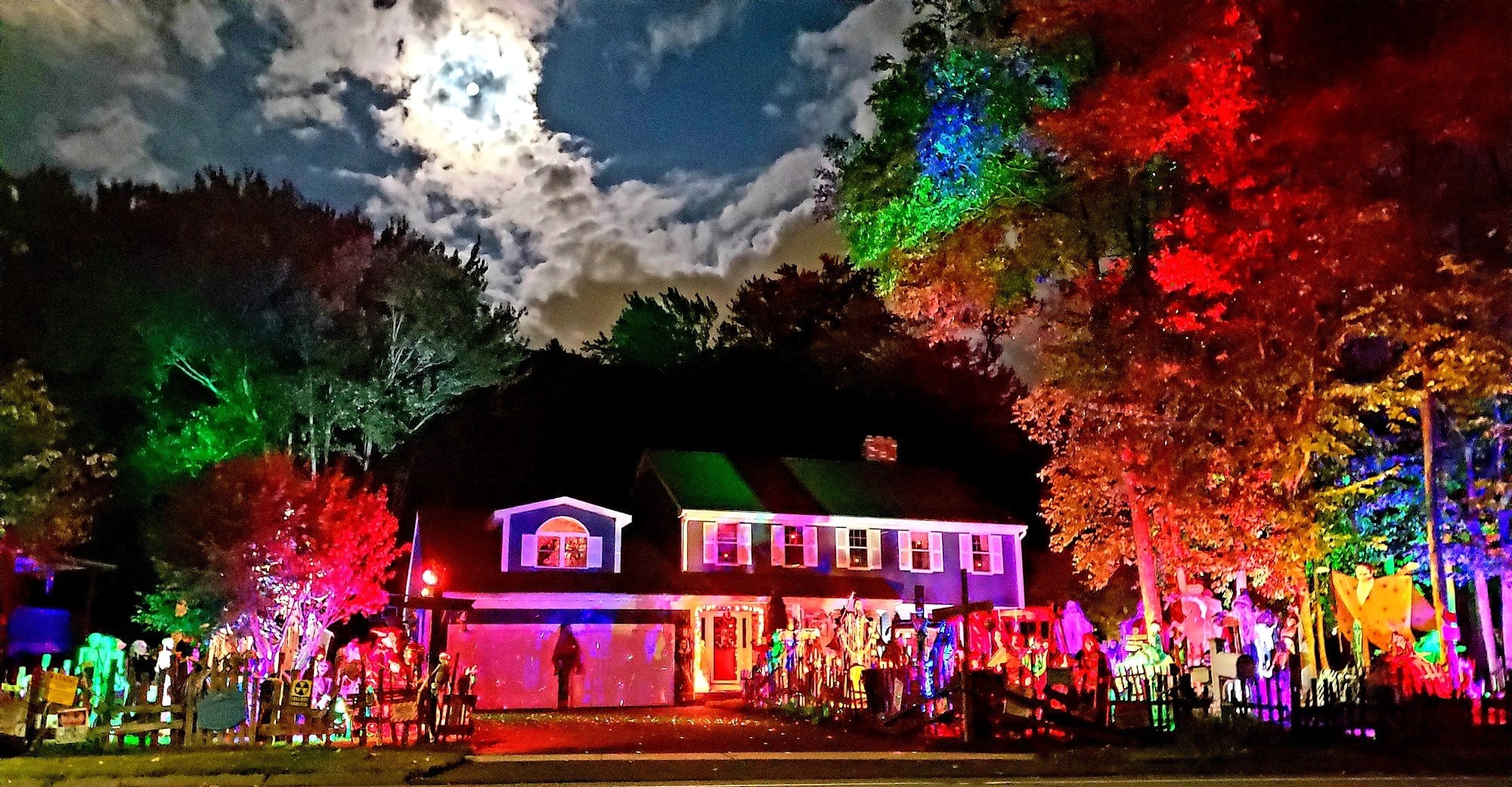 A house and trees lit red, green and orange at night under a cloudy full moon, with Halloween figures across the front yard