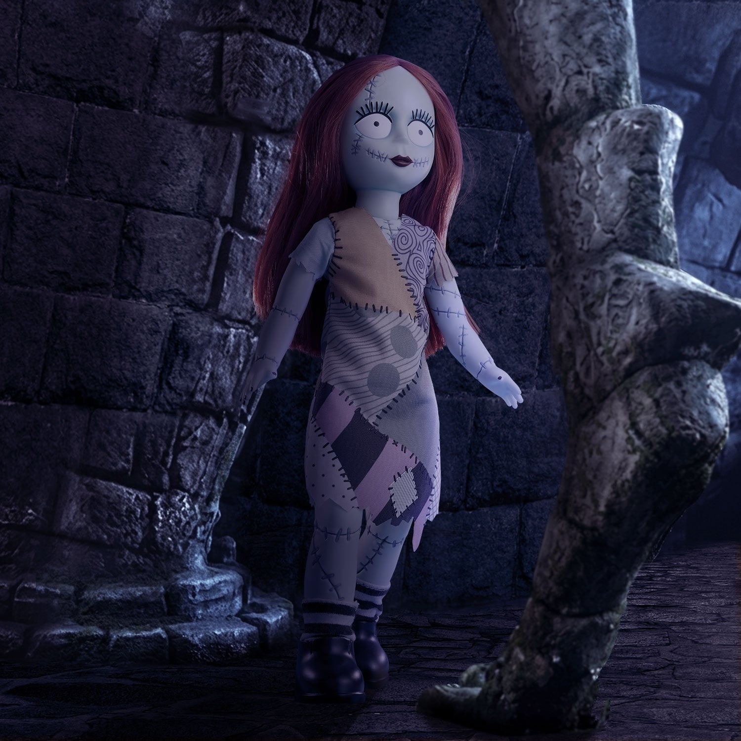 Mezco's LDD Presents Sally doll with long red hair and a patchwork dress, standing beside a stone wall