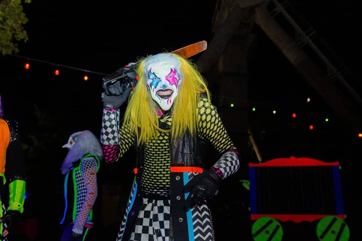 A clown with yellow hair and face paint in a checkered costume stands under string lights at night