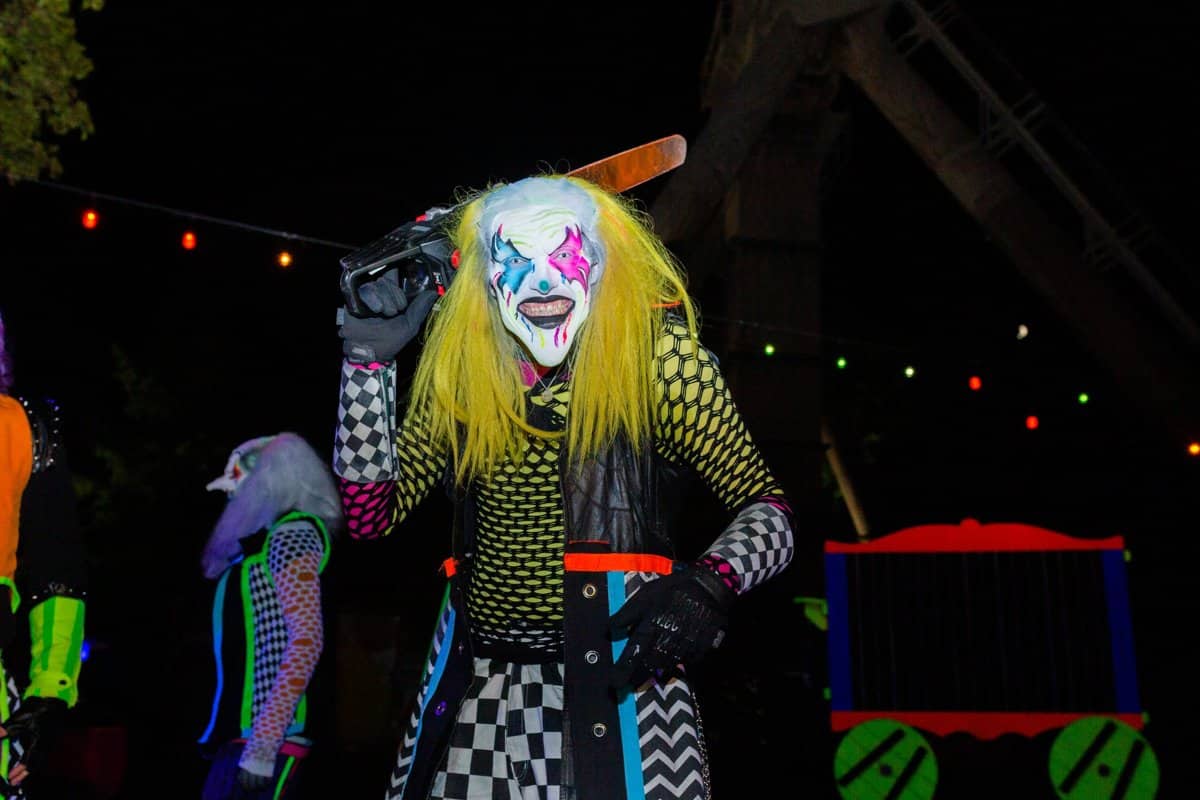 A clown with yellow hair and a painted face raises a bat in front of carnival lights at night