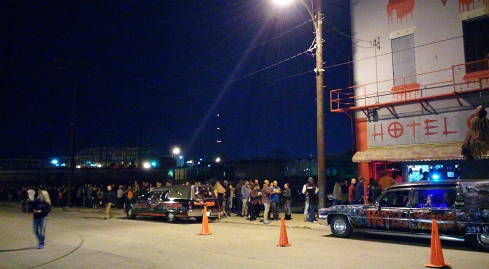 People line up at night outside a white building with a Hotel sign, with two painted hearses parked in front