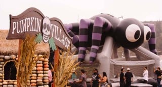 A carved wooden Pumpkin Village sign with a skull, beside a hut walled with stacked pumpkins and a giant purple and black striped spider bounce house