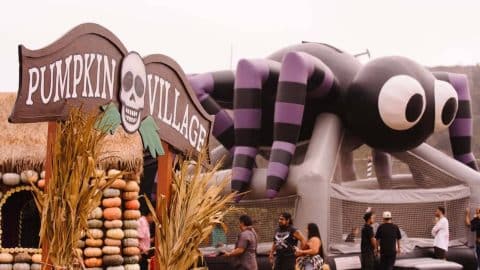A carved wooden Pumpkin Village sign with a skull, beside a hut walled with stacked pumpkins and a giant purple and black striped spider bounce house