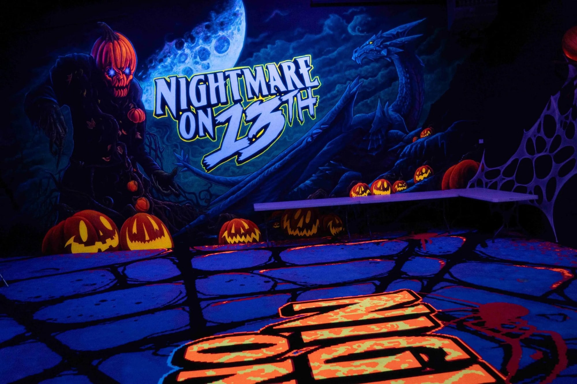 A blacklight mural of a pumpkin-headed figure, a full moon, a blue dragon, and glowing jack-o'-lanterns around the Nightmare on 13th logo