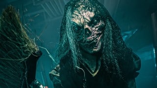 A Nightmare Wharf performer in a skull-like mask, veil and tattered dress reaches toward the camera in blue fog, holding a broom