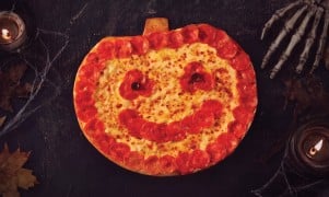 A pumpkin-shaped pizza ringed with pepperoni, with a pepperoni smile and olive eyes, on a dark table with candles, cobwebs and a skeleton hand