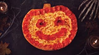A pumpkin-shaped pizza ringed with pepperoni, with a pepperoni smile and olive eyes, on a dark table with candles, cobwebs and a skeleton hand