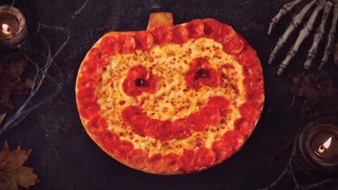 A pumpkin-shaped pizza ringed with pepperoni, with a pepperoni smile and olive eyes, on a dark table with candles, cobwebs and a skeleton hand