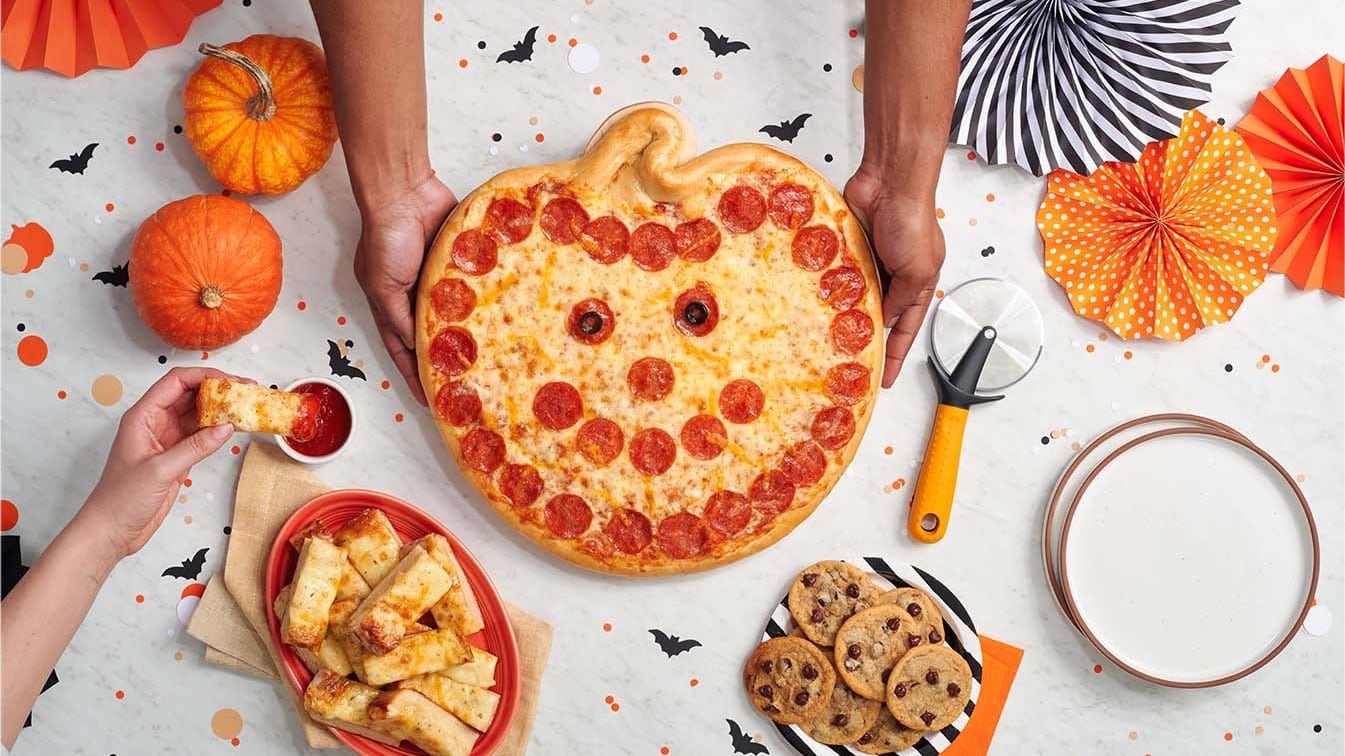 Two hands set a pumpkin-shaped pepperoni pizza with a smiling face on a table with small pumpkins, cookies and a pizza cutter