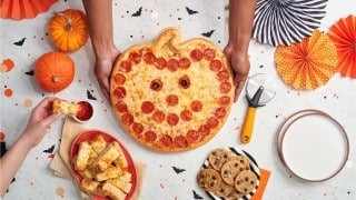 Two hands set a pumpkin-shaped pepperoni pizza with a pepperoni smile and olive eyes on a table beside pumpkins, cheese bread, and chocolate chip cookies