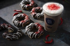 Paris Baguette's Ghost Pepper Chocolate Marble Mochi Donuts striped with white icing and red sugar, beside a Ghost Pepper Iced Hot Chocolate and red chili peppers