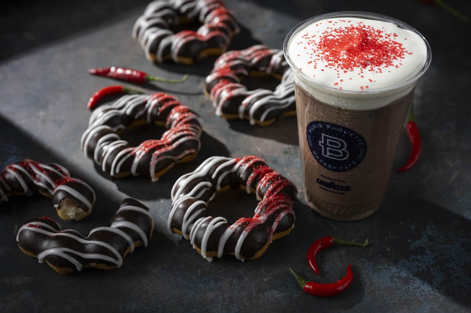 Paris Baguette's Ghost Pepper Chocolate Marble Mochi Donuts striped with white icing and red sugar, beside a Ghost Pepper Iced Hot Chocolate and red chili peppers