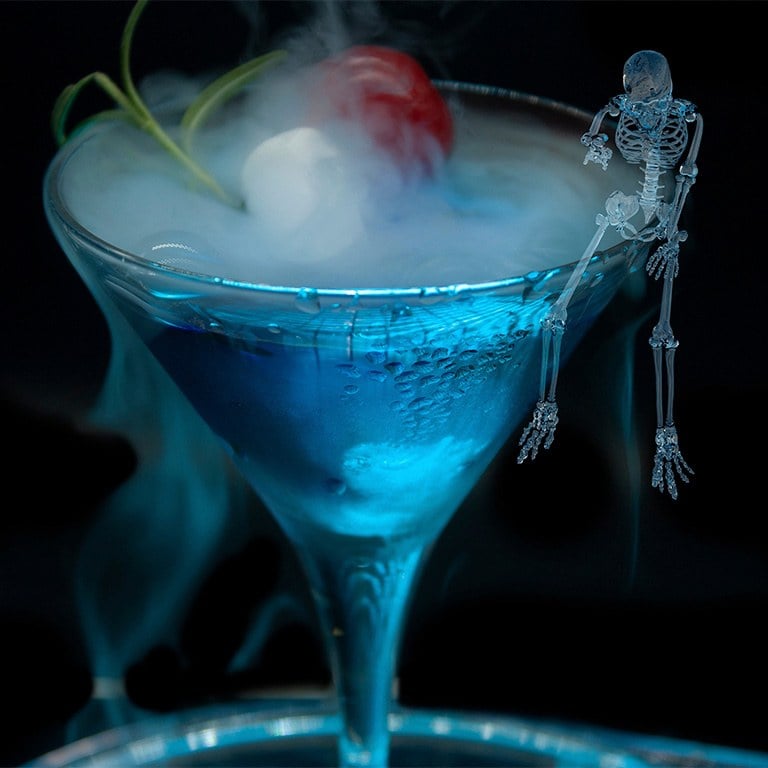 A blue cocktail in a coupe glass with smoke and a small skeleton perched on the rim