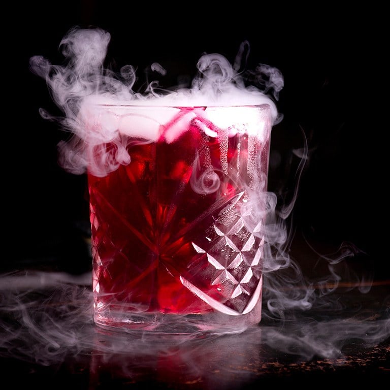 A red cocktail in a cut-glass tumbler with smoke pouring over the rim