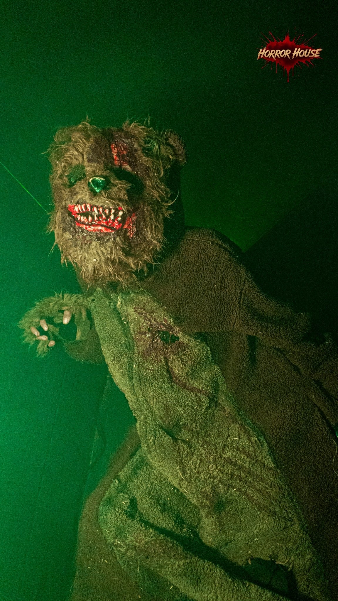 A performer in a matted teddy bear costume with bloody teeth reaches a clawed hand forward under green light