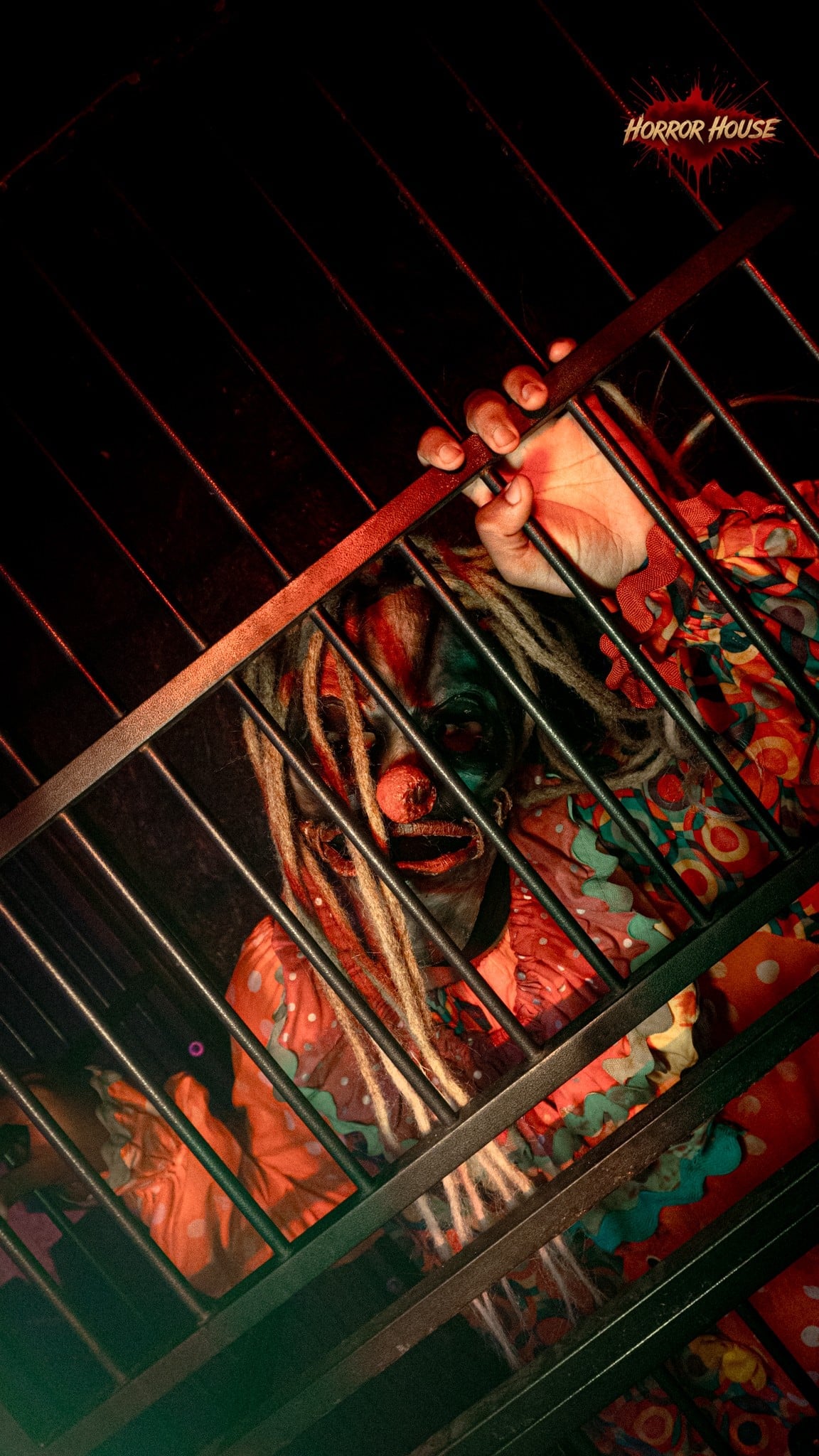 A clown in an orange polka-dot costume with long dreadlocks grips the bars of a cage and looks through them