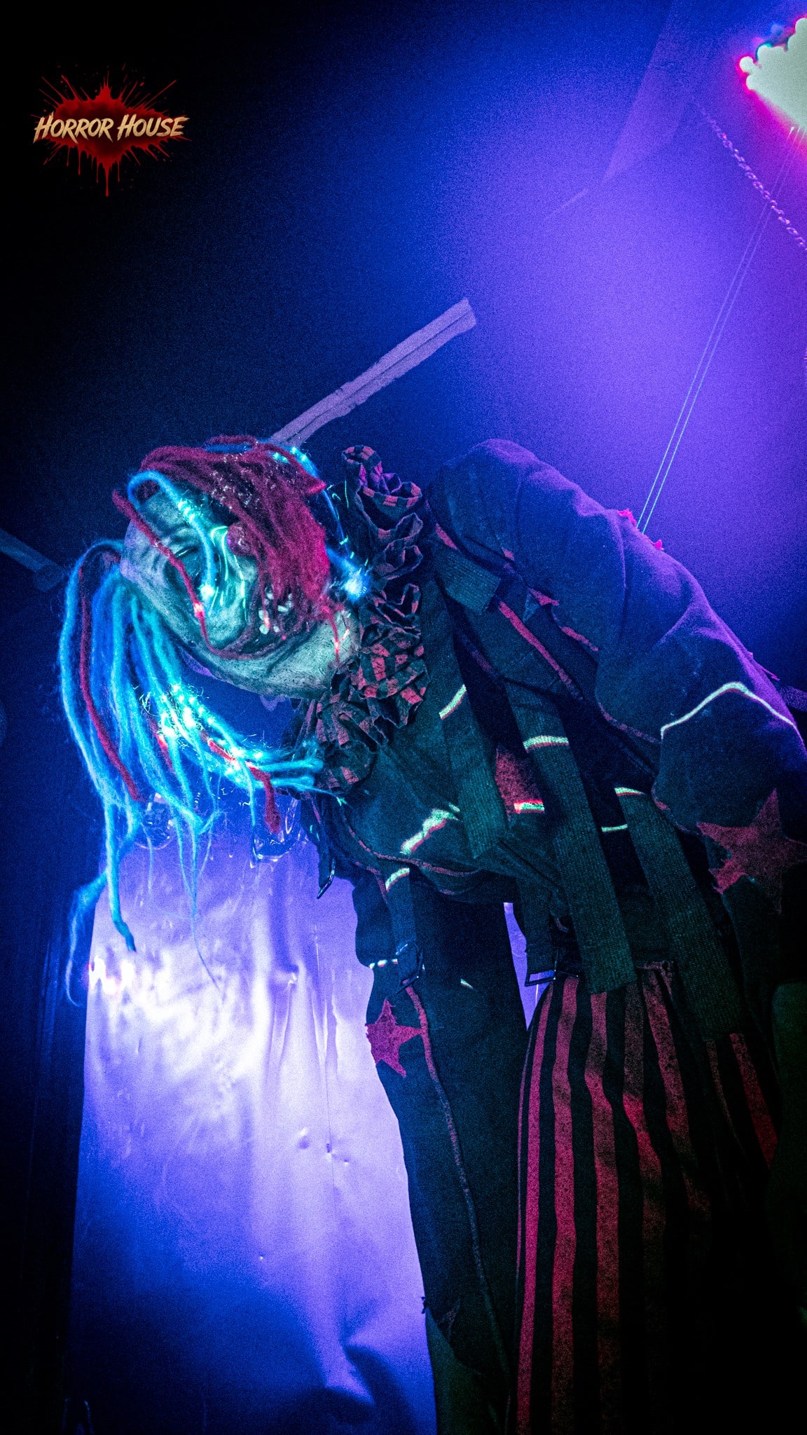 A clown with blue and red dreadlocks, a ruffled collar, and striped pants leans forward under purple light