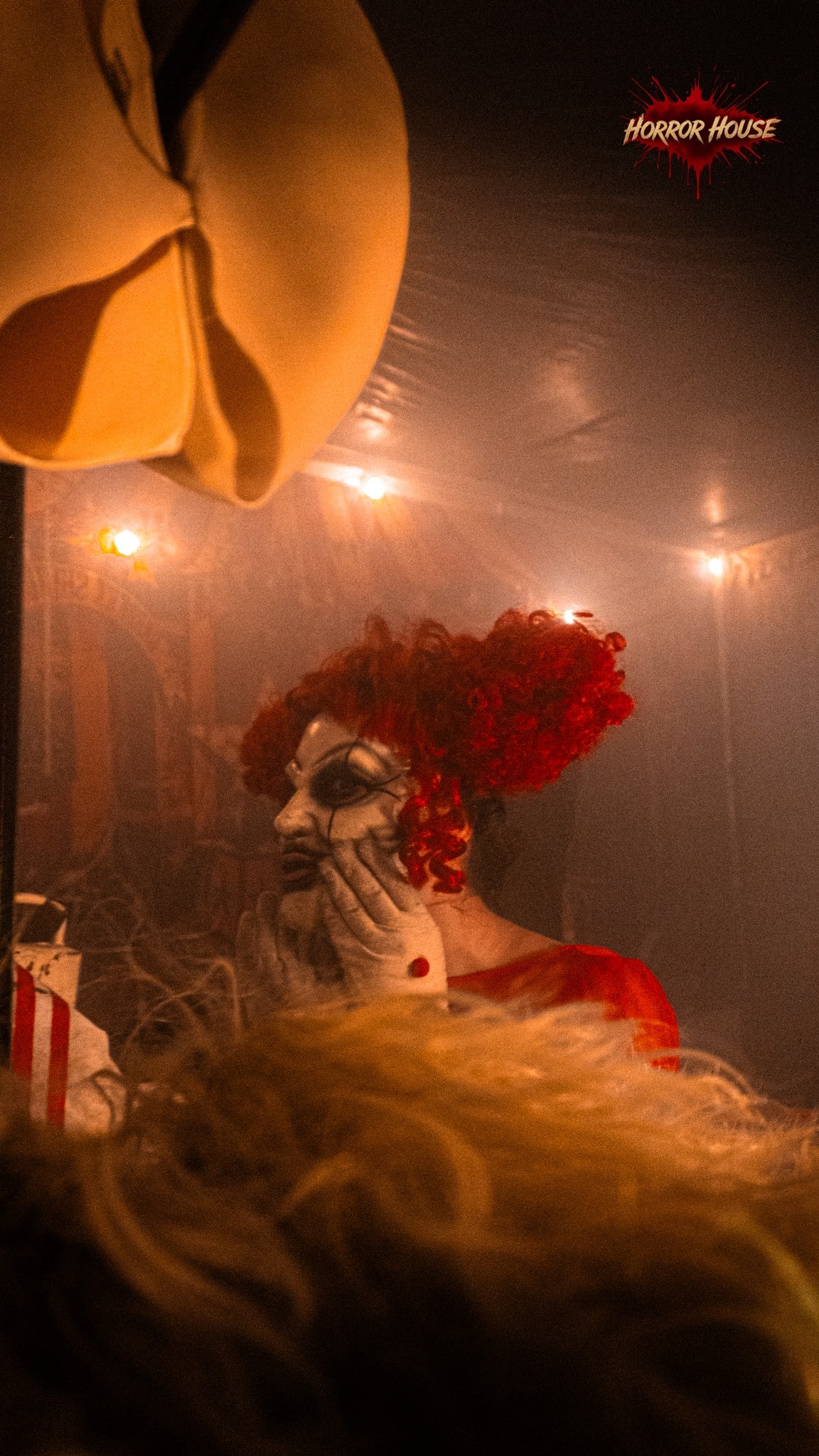 A clown with curly red hair, a white face, and white gloves holds its face in a hazy room lit by small bulbs