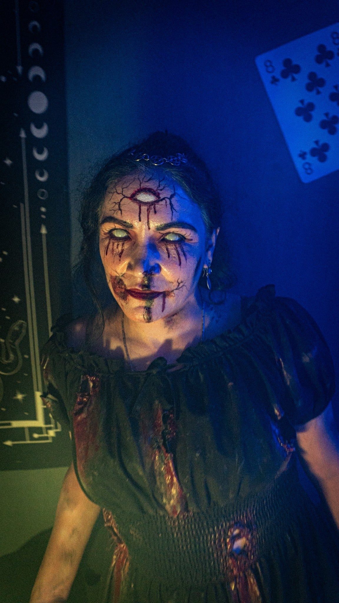 A performer with white eyes and a third eye painted on the forehead stands under blue light, with a moon-phase chart and a playing card on the wall