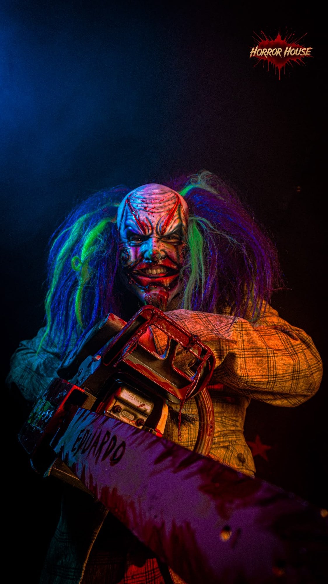 A clown with green and blue hair holds a chainsaw toward the camera