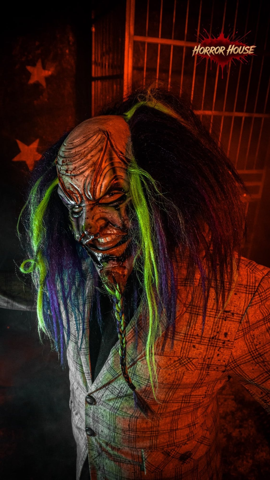 A clown with a scarred mask, a braided beard, green and purple hair, and a checked jacket stands in front of a cage and a red wall with stars