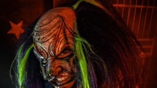 A clown in a scarred mask with long dark hair streaked green, in front of a red wall with a star and cage bars