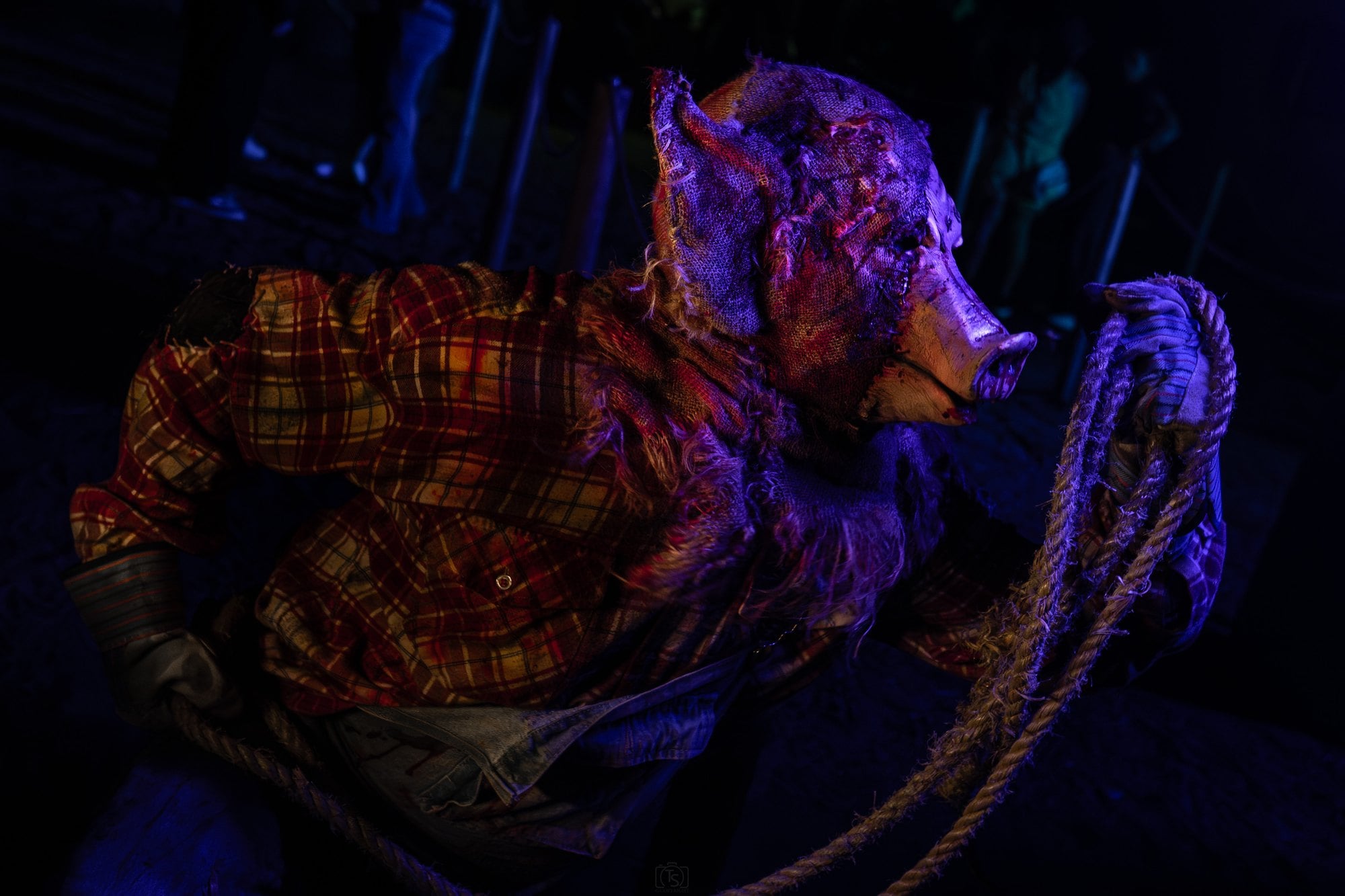 A figure in a pig mask, a burlap hood, and a plaid shirt holds a coiled rope under purple light