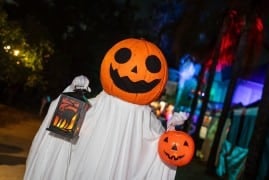 A person in a white sheet ghost costume with a jack-o'-lantern head holds a lit lantern and an orange plastic pumpkin pail outdoors at night