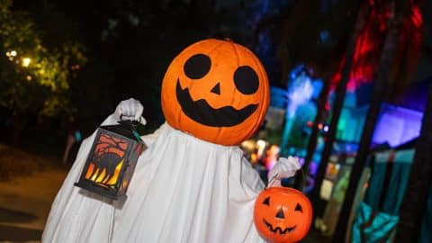 A person in a white sheet ghost costume with a jack-o'-lantern head holds a lit lantern and an orange plastic pumpkin pail outdoors at night