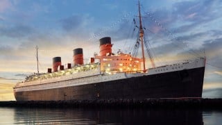 The Queen Mary ocean liner docked in Long Beach at dusk, with string lights running from its masts above three red and black funnels
