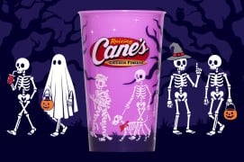 A purple cup with a red Raising Cane's logo, printed with skeletons and a skeleton dog in a red bandana, in front of a purple night scene with skeletons, a ghost, and jack-o'-lantern pails