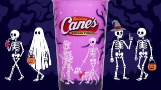 A purple cup with a red Raising Cane's logo, printed with skeletons and a skeleton dog in a red bandana, in front of a purple night scene with skeletons, a ghost, and jack-o'-lantern pails