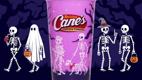 A purple cup with a red Raising Cane's logo, printed with skeletons and a skeleton dog in a red bandana, in front of a purple night scene with skeletons, a ghost, and jack-o'-lantern pails