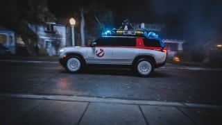 A white and red SUV with a ghost logo on its door and blue lights and gear on its roof drives down a dark street past a lit street lamp