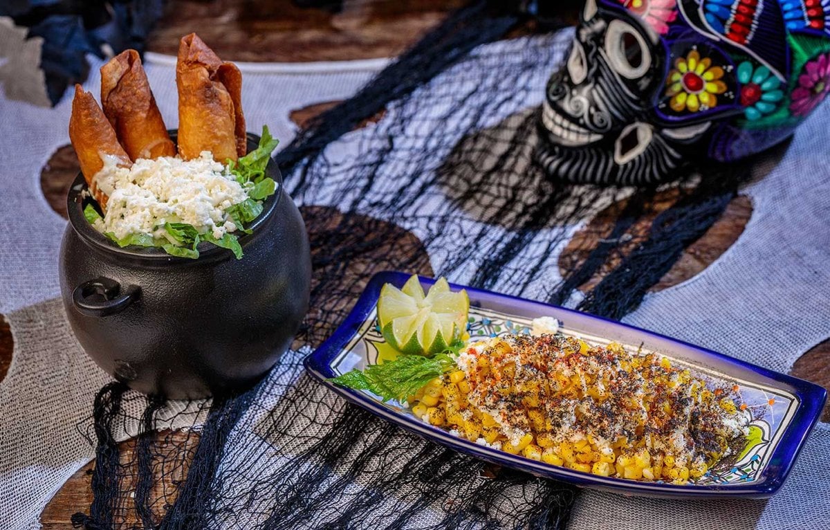 Rolled flautas in a mini black cauldron beside a plate of seasoned street corn