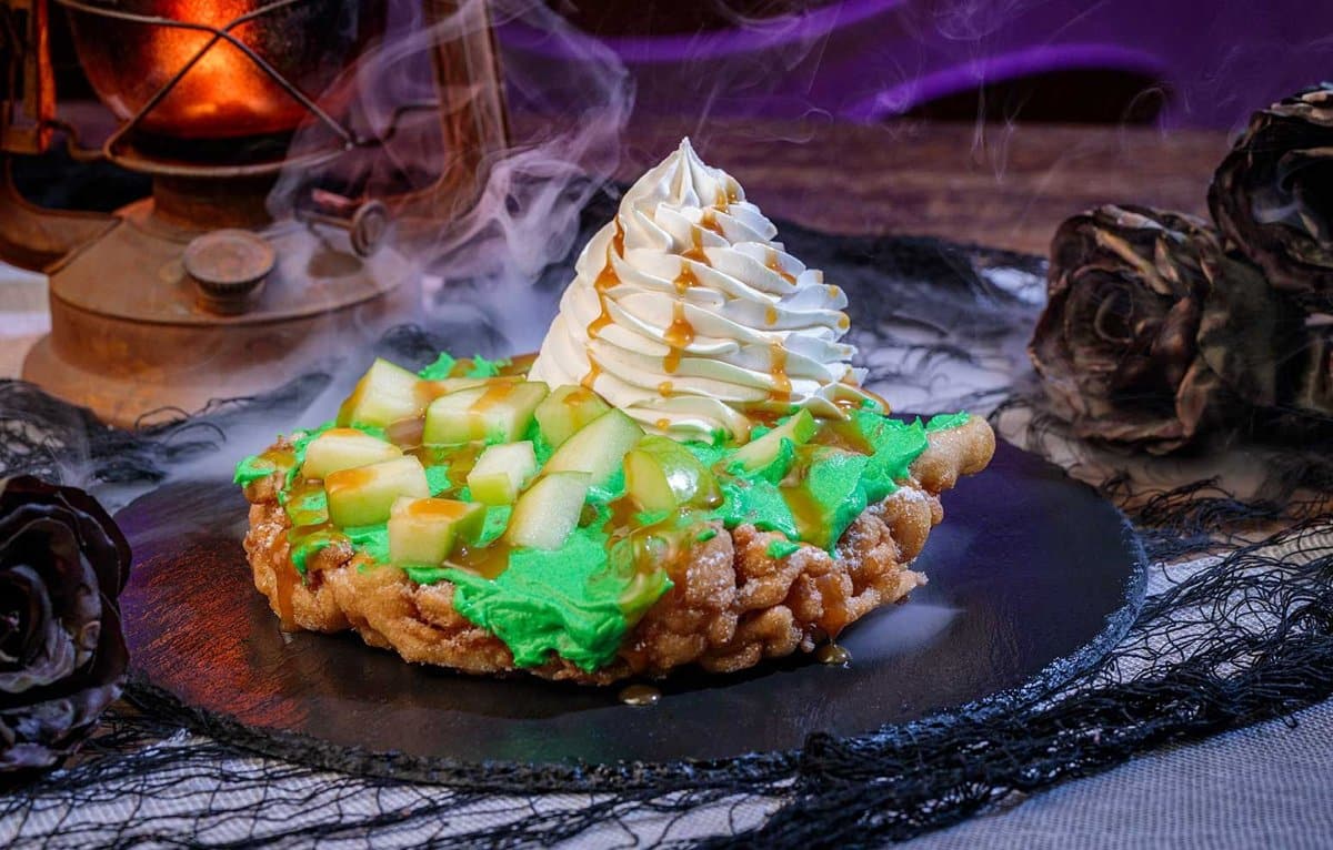 A funnel cake topped with green icing, diced apples, caramel, and whipped cream