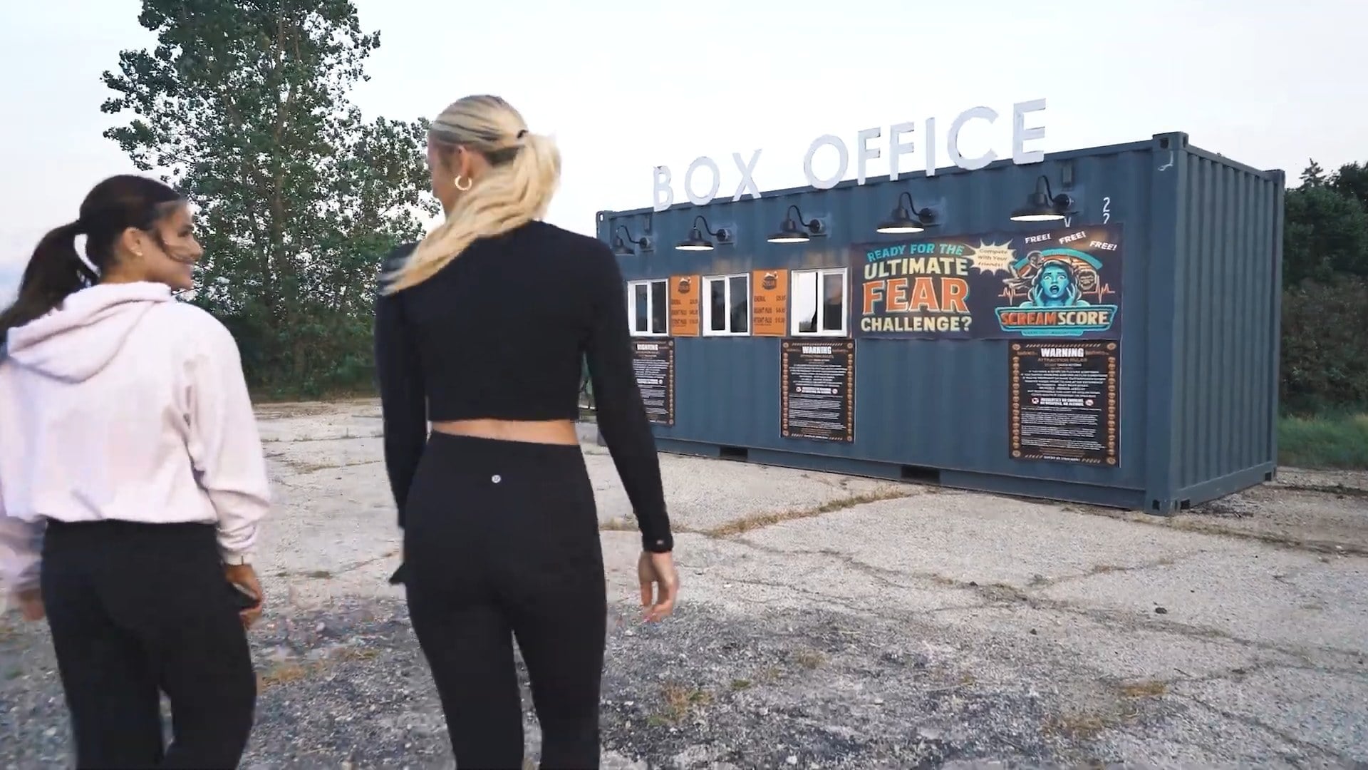 Two people walk toward a blue shipping-container box office with a Scream Score banner reading Ready for the Ultimate Fear Challenge