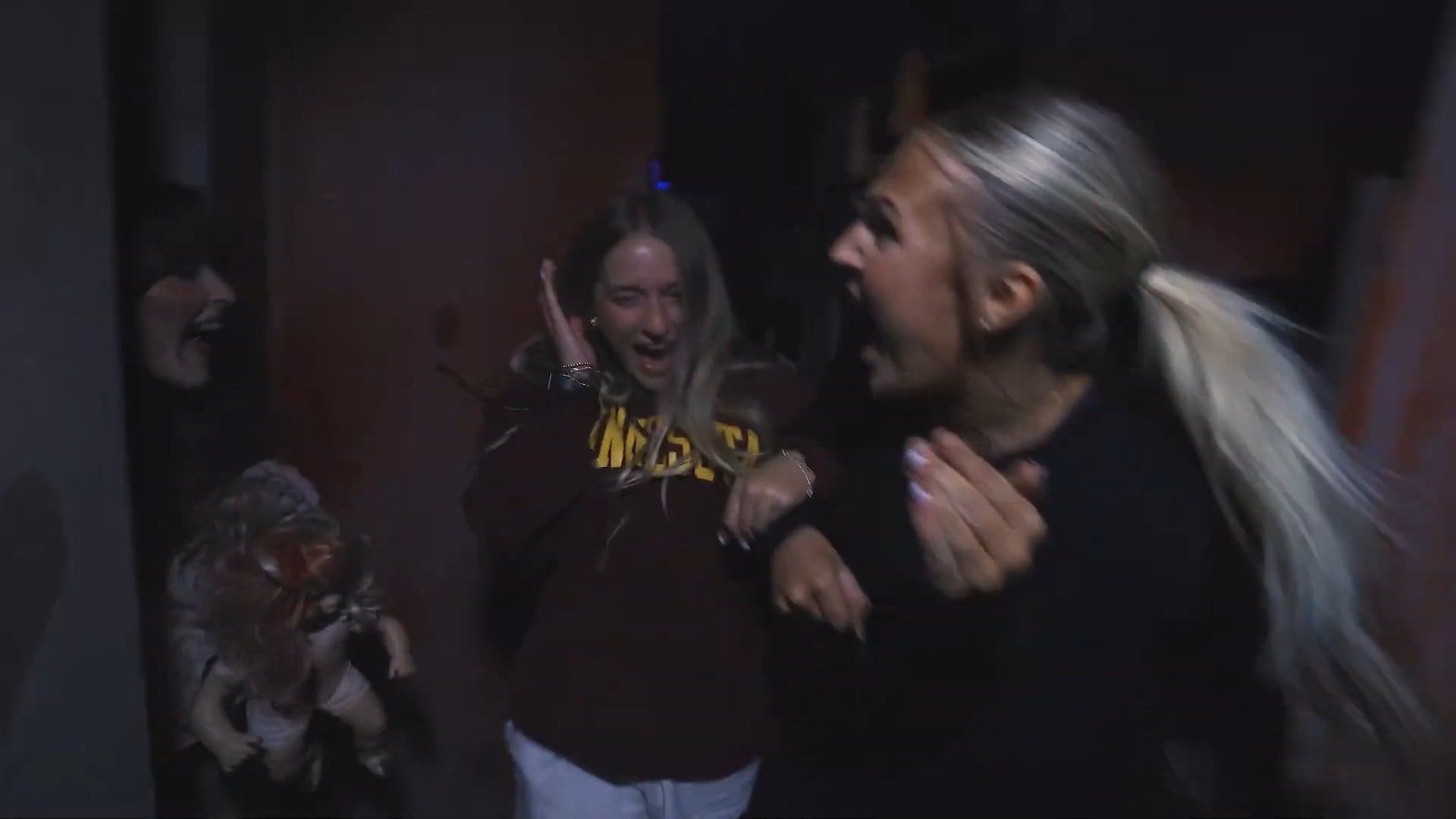 Three people react in a dark room of a haunted house, one with her eyes shut