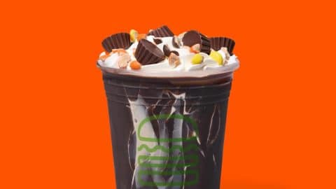 Shake Shack Reese's Peanut Butter Cup Shake in a crackable dark chocolate shell, topped with whipped cream, Reese's Mini Peanut Butter Cups, and chopped Reese's Pieces, on an orange background