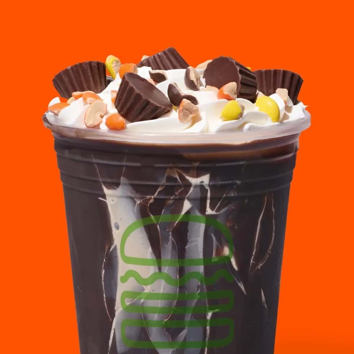 Shake Shack Reese's Peanut Butter Cup Shake: a dark chocolate shell coats the inside of the cup, topped with whipped cream, Reese's Mini Peanut Butter Cups, and chopped Reese's Pieces, on an orange background