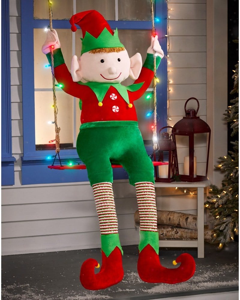 An elf in a green suit and striped stockings sitting on a swing hung with lights