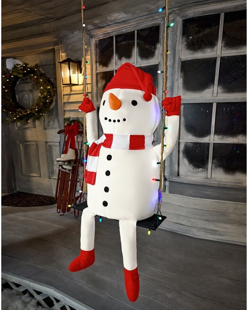 A snowman in a red hat and scarf sitting on a swing hung with lights