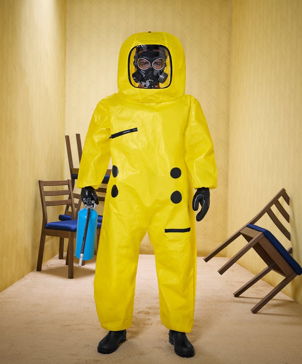 A person in a yellow hazmat suit and gas mask stands in a yellow room with wooden chairs