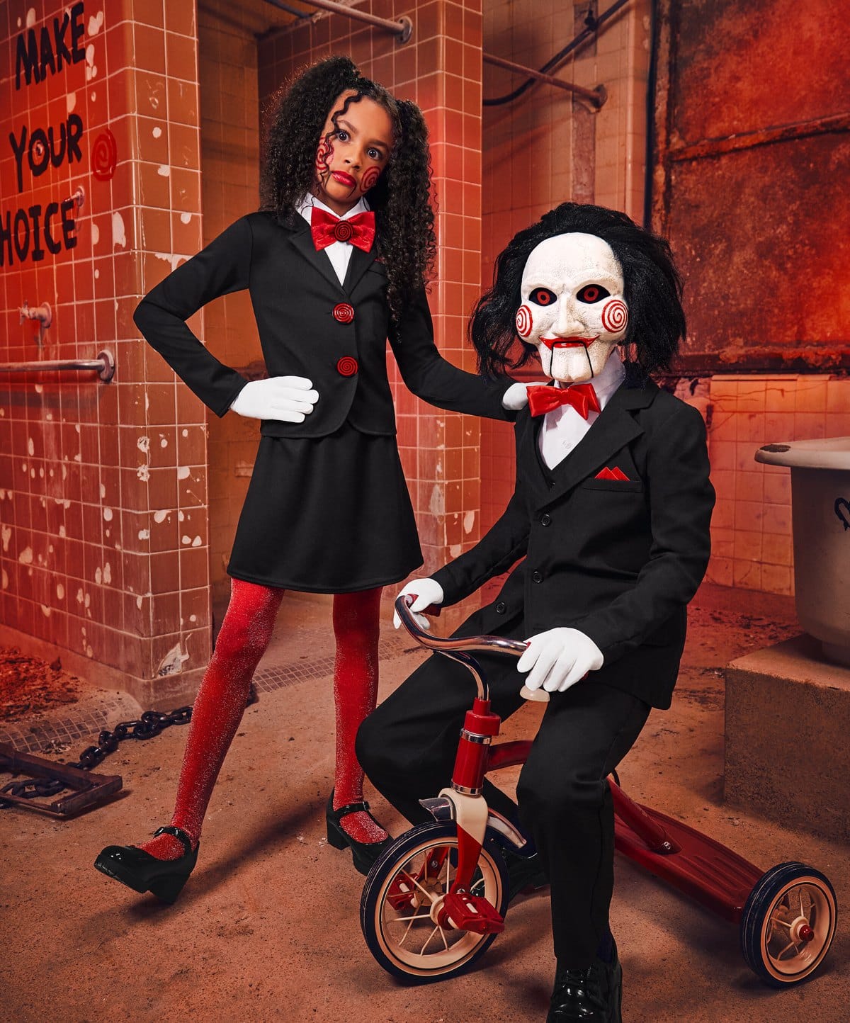 Two kids in Saw costumes, one in a black suit dress and one as Billy the Puppet on a red tricycle