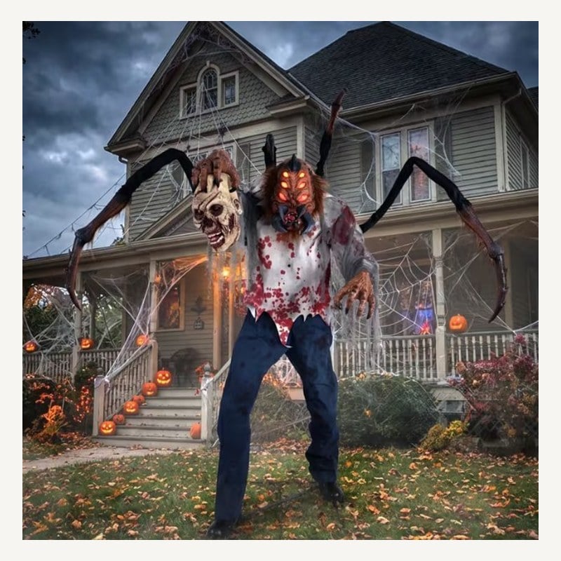 A life-size spider-legged monster in a bloody shirt holding a severed head in front of a decorated house