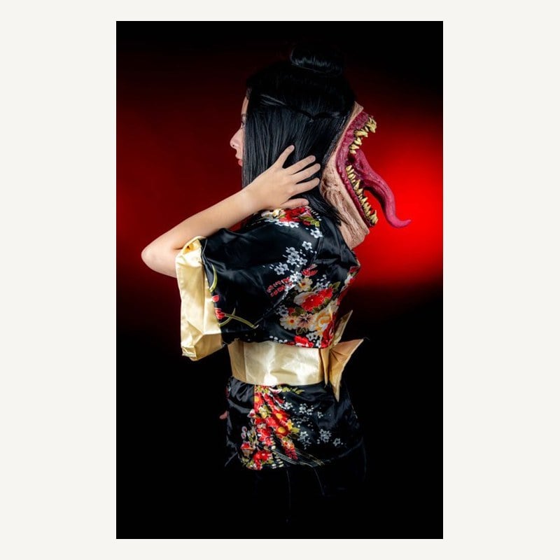 A woman seen from behind wearing a mask of a giant fanged mouth with a long tongue on the back of her head