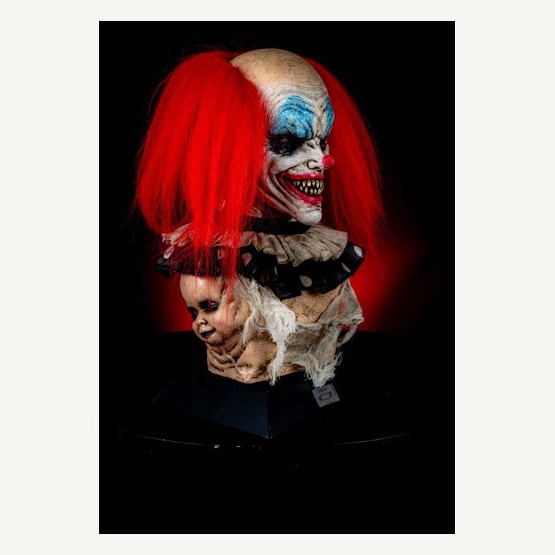 A clown bust with red hair, blue eyeshadow, and sharp yellow teeth, with pale baby heads at its base