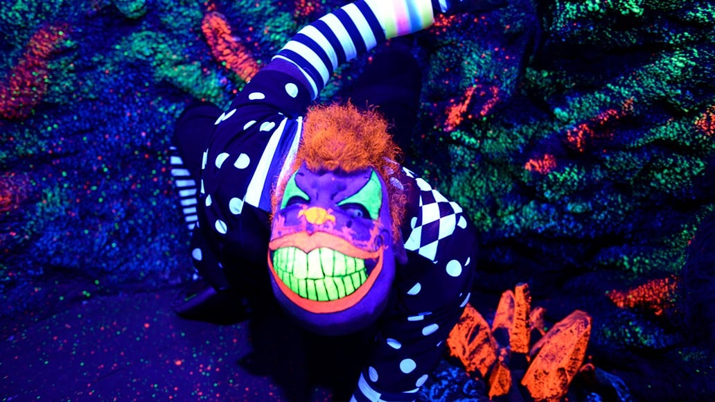 A performer in a clown costume with glowing orange hair and green teeth leans toward the camera in a room of blacklight paint